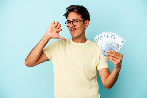 Hombre joven de raza mixta con billetes aislados sobre fondo azul se siente orgulloso y seguro de sí mismo, ejemplo a seguir.