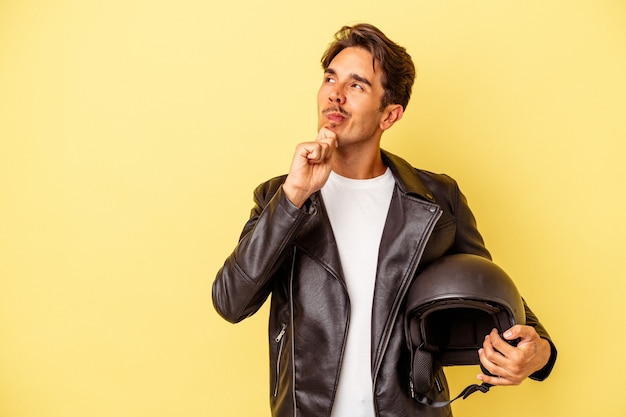 Hombre joven de raza mixta con casco aislado sobre fondo amarillo mirando hacia los lados con expresión dudosa y escéptica.