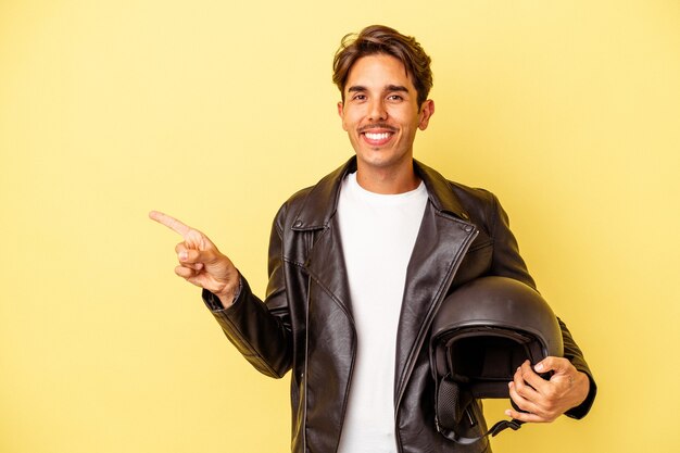 Hombre joven de raza mixta con casco aislado sobre fondo amarillo sonriendo y apuntando a un lado, mostrando algo en el espacio en blanco.