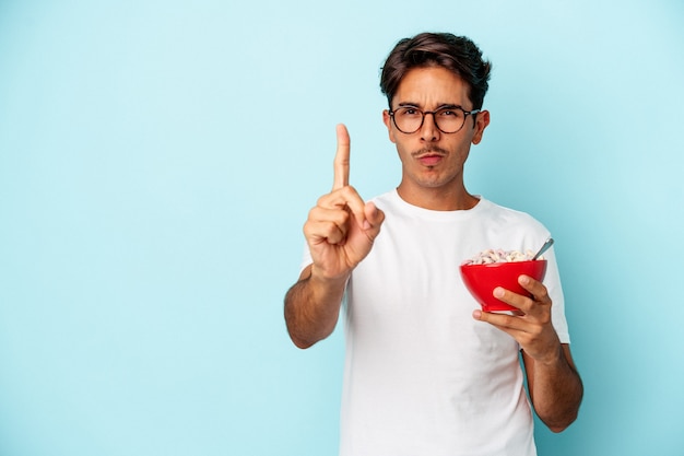 Hombre joven de raza mixta con cereales aislado sobre fondo azul que muestra el número uno con el dedo.