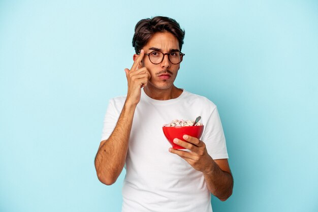 Hombre joven de raza mixta con cereales aislados sobre fondo azul apuntando la sien con el dedo, pensando, centrado en una tarea.