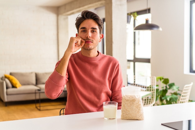 Hombre joven de raza mixta comiendo avena y leche para el desayuno en su cocina con los dedos en los labios guardando un secreto.