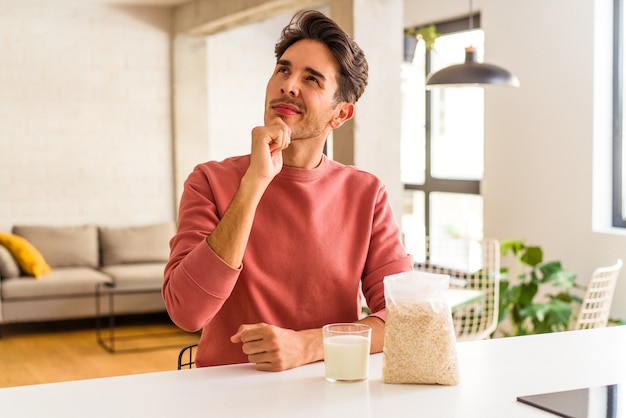 Hombre joven de raza mixta comiendo avena y leche para el desayuno en su cocina mirando hacia los lados con expresión dudosa y escéptica.