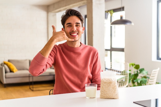 Hombre joven de raza mixta comiendo avena y leche para el desayuno en su cocina mostrando un gesto de llamada de teléfono móvil con los dedos.