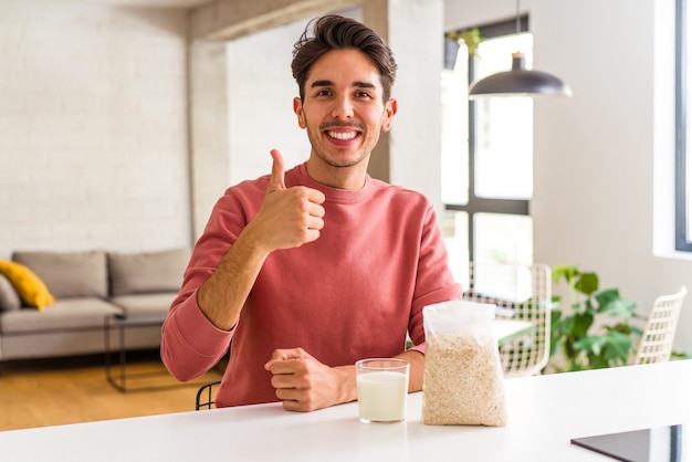 Hombre joven de raza mixta comiendo avena y leche para el desayuno en su cocina sonriendo y levantando el pulgar hacia arriba