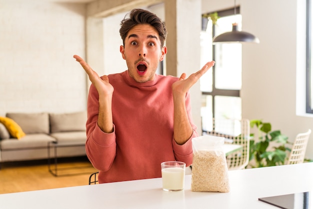 Hombre joven de raza mixta comiendo avena y leche para el desayuno en su cocina sorprendido y conmocionado.