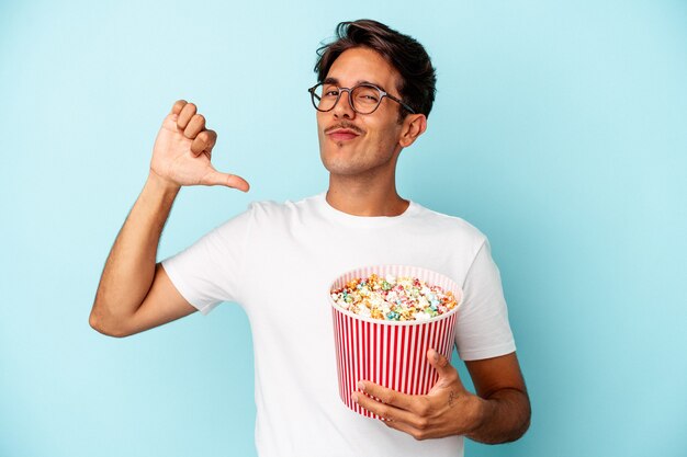 Hombre joven de raza mixta comiendo palomitas de maíz aisladas sobre fondo azul se siente orgulloso y seguro de sí mismo, ejemplo a seguir.