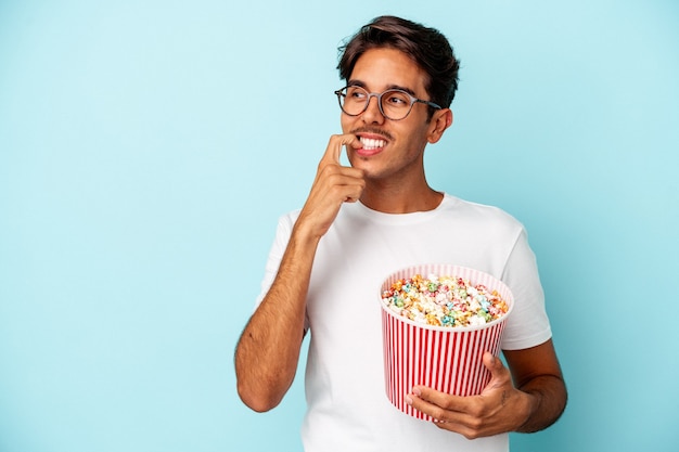 Hombre joven de raza mixta comiendo palomitas de maíz aislado sobre fondo azul relajado pensando en algo mirando un espacio de copia.