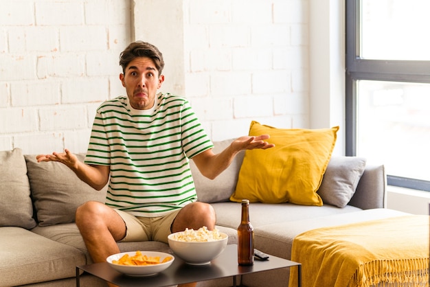 Hombre joven de raza mixta comiendo palomitas de maíz sentado en el sofá se encoge de hombros y abre los ojos confundidos.