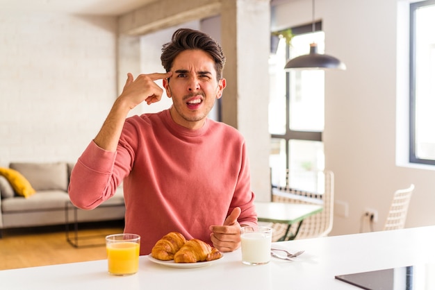Hombre joven de raza mixta desayunando en una cocina por la mañana mostrando un gesto de decepción con el dedo índice.