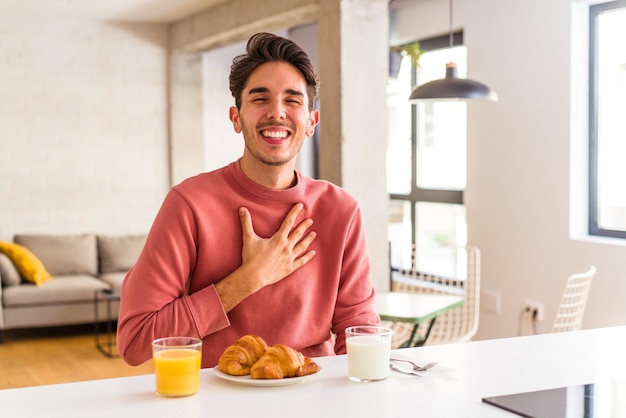 Foto hombre joven de raza mixta desayunando en una cocina en la mañana se ríe a carcajadas manteniendo la mano en el pecho.