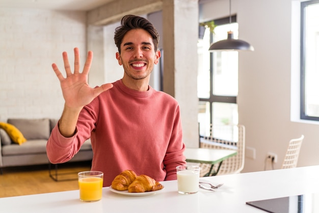 Hombre joven de raza mixta desayunando en una cocina en la mañana sonriendo alegre mostrando el número cinco con los dedos.