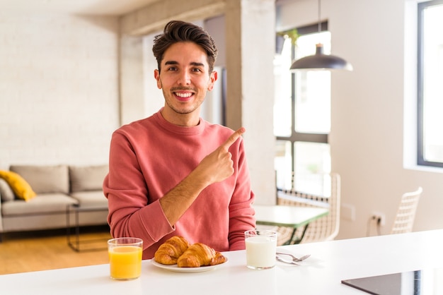 Foto hombre joven de raza mixta desayunando en una cocina por la mañana sonriendo y apuntando a un lado, mostrando algo en el espacio en blanco.