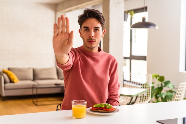 Hombre joven de raza mixta desayunando en su cocina de pie con la mano extendida mostrando la señal de stop, impidiéndote.