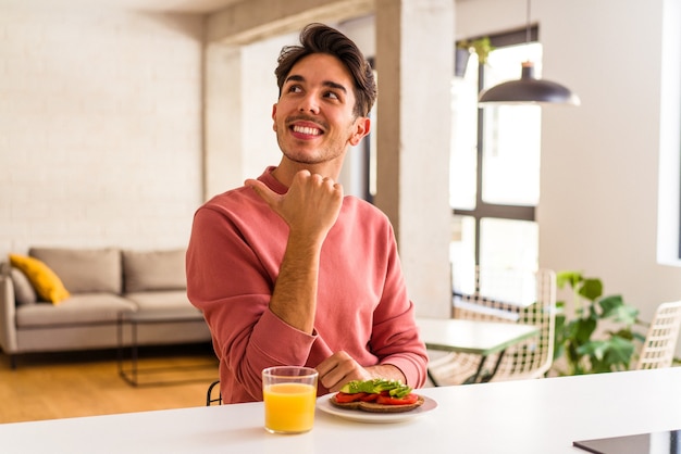 Foto hombre joven de raza mixta desayunando en su cocina señala con el dedo pulgar, riendo y despreocupado.