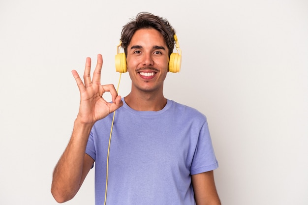 Hombre joven de raza mixta escuchando música aislada sobre fondo azul alegre y confiado mostrando gesto ok.