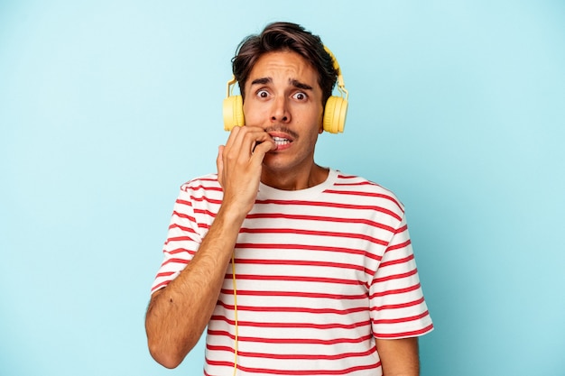 Hombre joven de raza mixta escuchando música aislada sobre fondo azul mordiéndose las uñas, nervioso y muy ansioso.