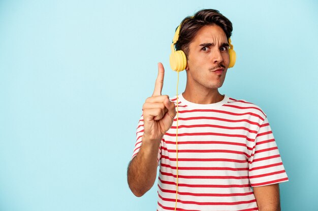 Hombre joven de raza mixta escuchando música aislado sobre fondo azul mostrando el número uno con el dedo.