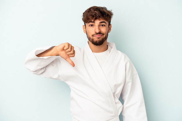 Hombre joven de raza mixta haciendo karate aislado sobre fondo azul mostrando un gesto de aversión, pulgares hacia abajo. Concepto de desacuerdo.