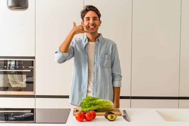 Hombre joven de raza mixta preparando una ensalada para el almuerzo mostrando un gesto de llamada de teléfono móvil con los dedos.