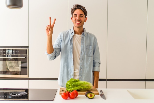 Hombre joven de raza mixta preparando una ensalada para el almuerzo mostrando el número dos con los dedos.