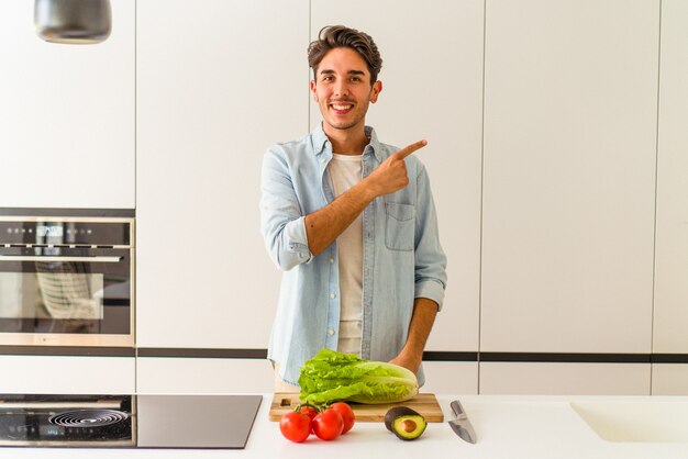 Foto hombre joven de raza mixta preparando una ensalada para el almuerzo sonriendo y apuntando a un lado, mostrando algo en el espacio en blanco.
