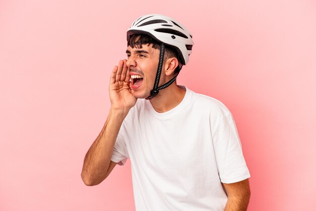 Hombre joven de raza mixta que llevaba un casco de bicicleta aislado sobre fondo rosa gritando y sosteniendo la palma cerca de la boca abierta.