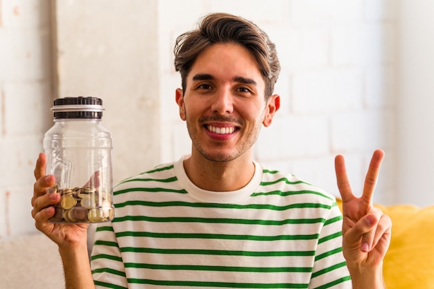 Hombre joven de raza mixta que sostiene la hucha en su sala de estar mostrando el número dos con los dedos.