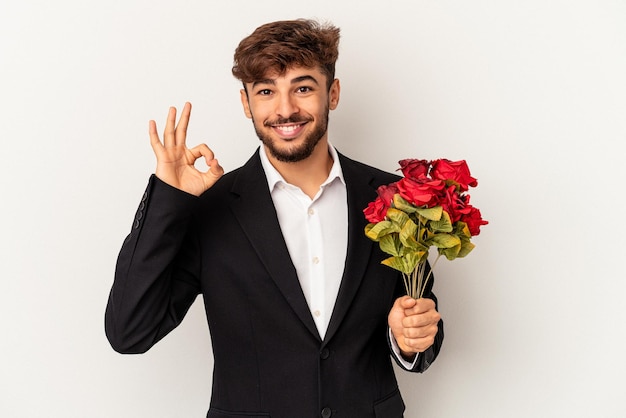 Hombre joven de raza mixta con ramo de rosas aislado sobre fondo blanco alegre y confiado mostrando gesto ok.