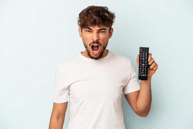 Hombre joven de raza mixta sosteniendo un controlador de tv aislado sobre fondo azul gritando muy enojado y agresivo.