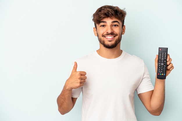 Hombre joven de raza mixta sosteniendo un controlador de tv aislado sobre fondo azul sonriendo y levantando el pulgar hacia arriba