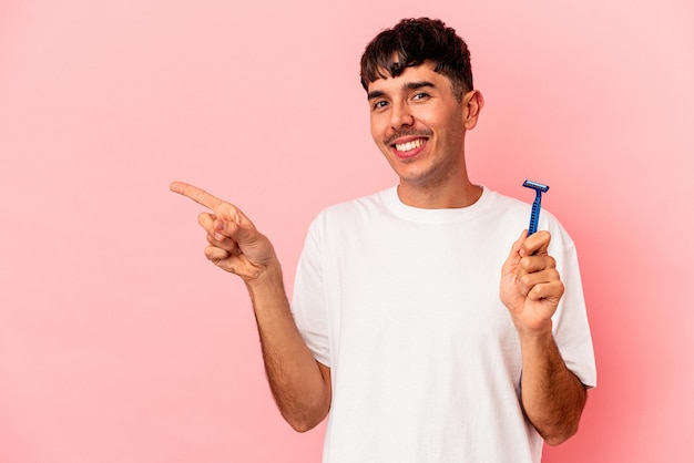 Hombre joven de raza mixta sosteniendo una hoja de afeitar aislada sobre fondo rosa sonriendo y apuntando a un lado, mostrando algo en el espacio en blanco.