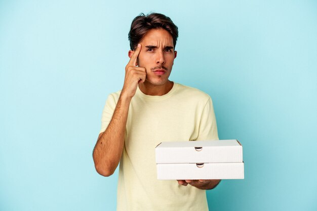 Hombre joven de raza mixta sosteniendo pizzas aisladas sobre fondo azul apuntando al templo con el dedo, pensando, centrado en una tarea.