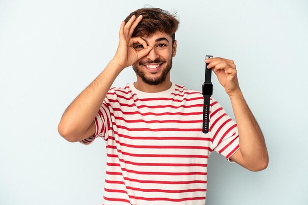 Hombre joven de raza mixta sosteniendo un reloj de pulsera aislado sobre fondo azul emocionado manteniendo el gesto ok en el ojo.