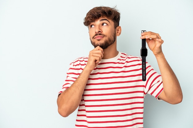 Hombre joven de raza mixta sosteniendo un reloj de pulsera aislado sobre fondo azul mirando hacia los lados con expresión dudosa y escéptica.