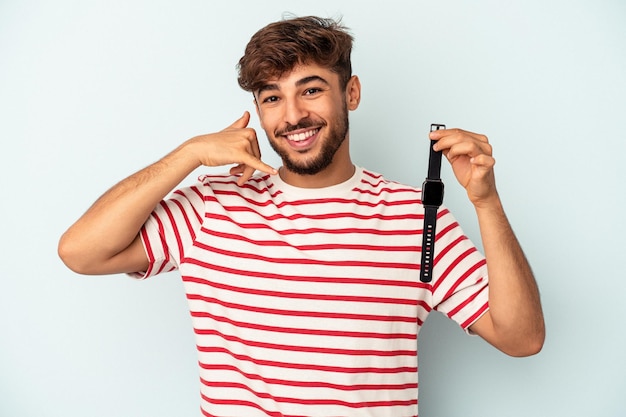 Hombre joven de raza mixta sosteniendo un reloj de pulsera aislado sobre fondo azul que muestra un gesto de llamada de teléfono móvil con los dedos.