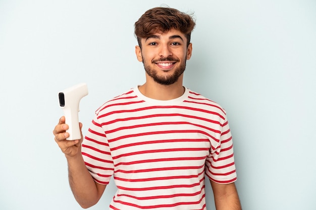 Hombre joven de raza mixta con termómetro aislado sobre fondo azul feliz, sonriente y alegre.