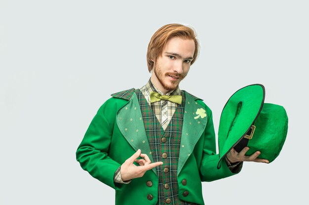 Foto el hombre joven del readhead en traje verde sostiene el sombrero en la mano y señala en él. guy mira en serio. aislado en gris