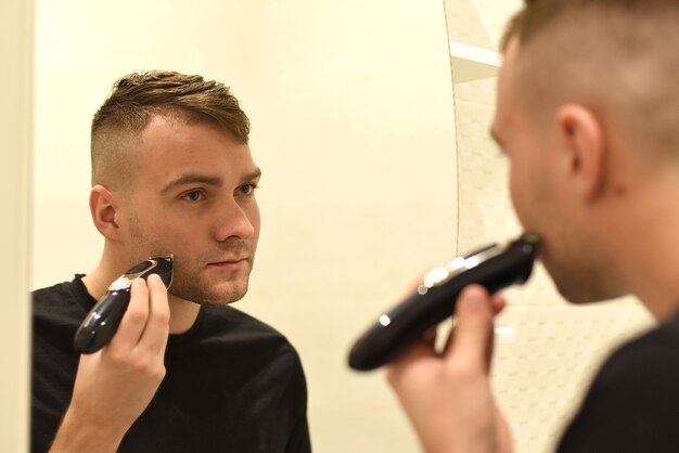 Hombre joven recortando la barba y mirando en el espejo en un baño