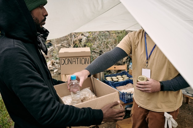 Hombre joven refugiado que sostiene la caja mientras el voluntario pone allí comida y agua