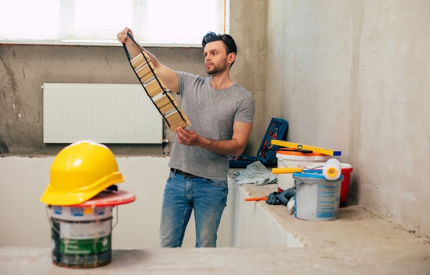 Hombre joven de reparación de viviendas o trabajador ocupado con la renovación de la casa, sostiene equipos de construcción, viste ropa de trabajo informal en la nueva construcción