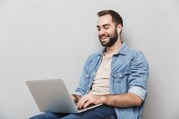 Foto hombre joven riendo con barba escribiendo portátil y usando earpod mientras está sentado en una silla aislada sobre pared gris