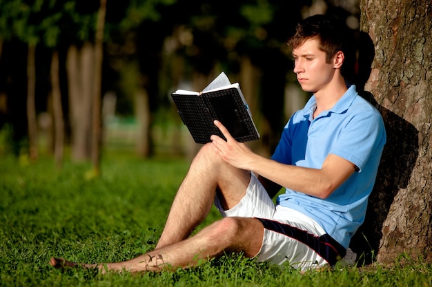 Hombre joven en ropa casual sentado en la hierba verde cerca de un árbol y un libro de lectura en el parque en un día claro de verano. Libertad interior y concepto de estilo de vida feliz