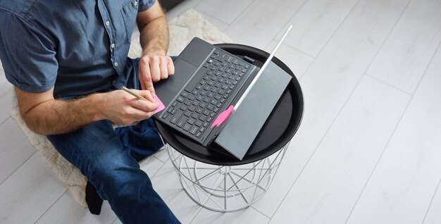 Hombre joven en ropa casual sentado en el piso y trabajando en su computadora portátil en casa. Quédese en casa y trabaje en línea