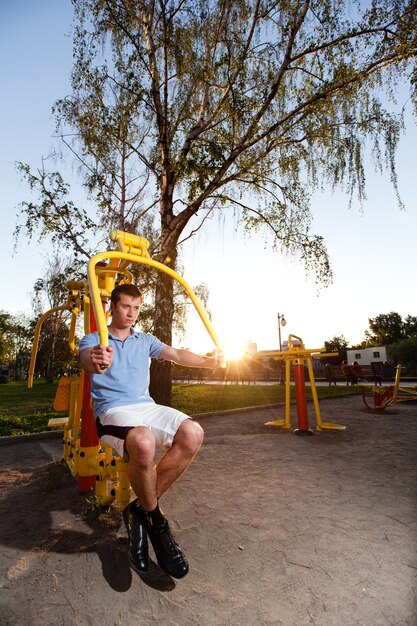 Hombre joven en ropa casual sentado y trabajando en la máquina de fitness al aire libre