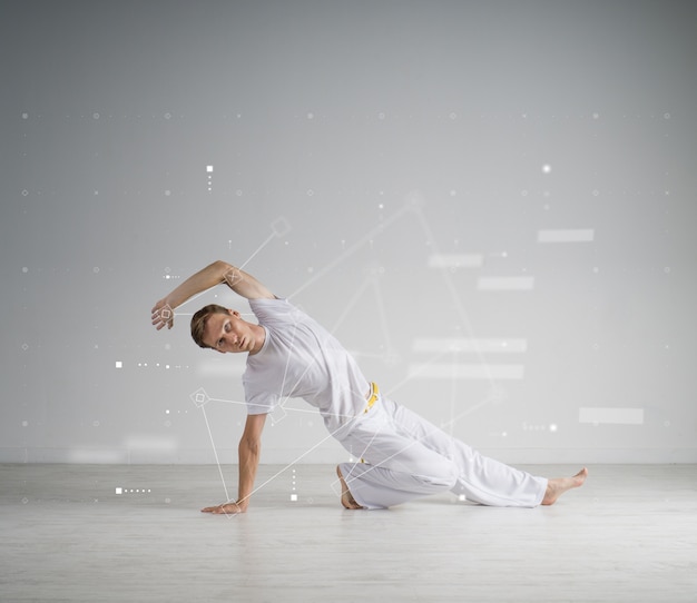 Hombre joven en ropa deportiva blanca realizando una patada. Entrenamiento de artes marciales bajo techo, capoeira.