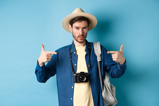 Hombre joven seguro y listo apuntando a sí mismo, autopromoción o presumido, yendo de vacaciones de verano, con sombrero de paja, sosteniendo una mochila con cámara, fondo azul.