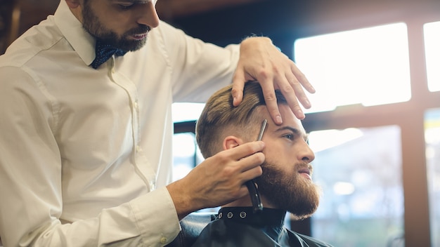 Hombre joven sentado en una barbería mientras peluquero trabaja con navaja