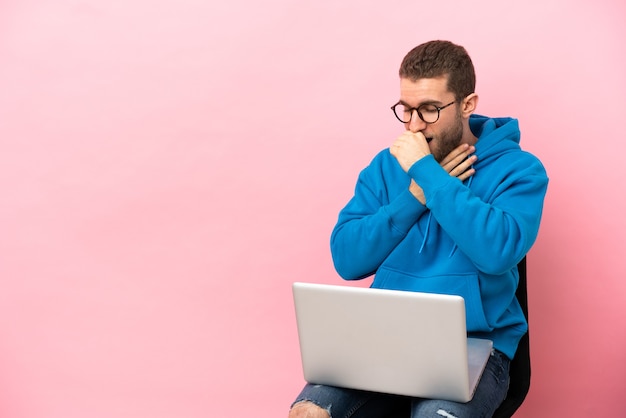 Un hombre joven sentado en una silla con una computadora portátil sufre de tos y se siente mal