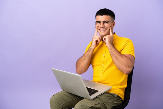 Hombre joven sentado en una silla con laptop sonriendo con una expresión feliz y agradable